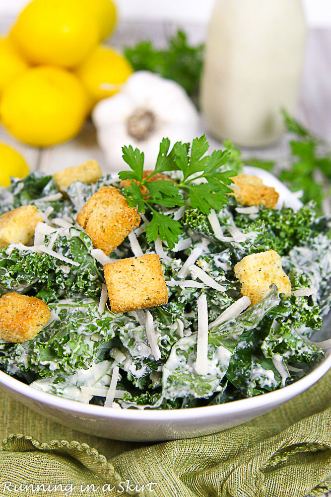 Kale Caesar Salad in a white bowl with lemon and garlic in the background.